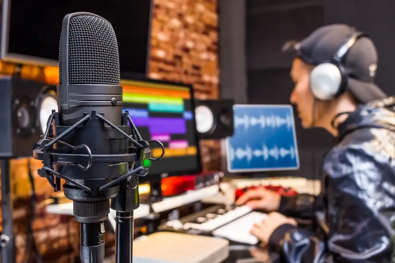 Micrófono profesional en un estudio de grabación con una persona trabajando en software de edición de audio al fondo.