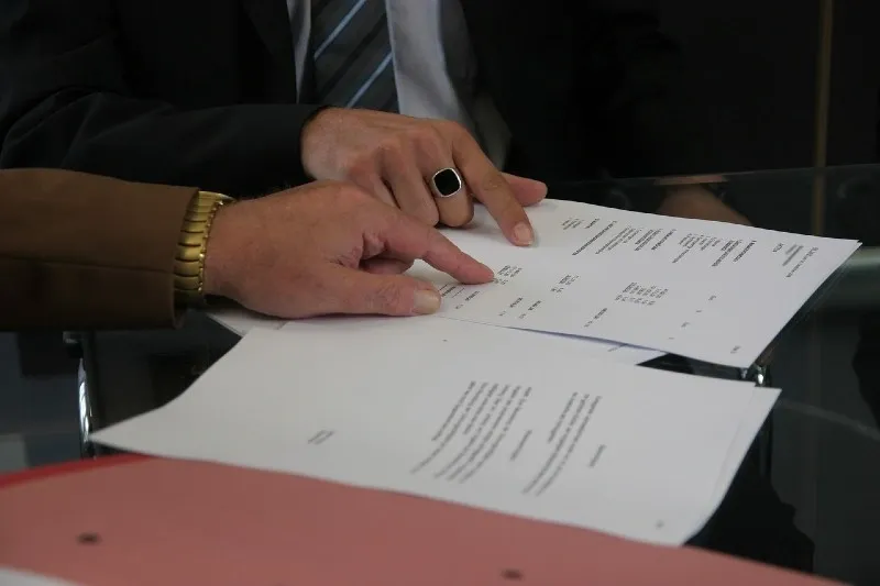 Two people in business attire reviewing and pointing at contract documents on a glass table.